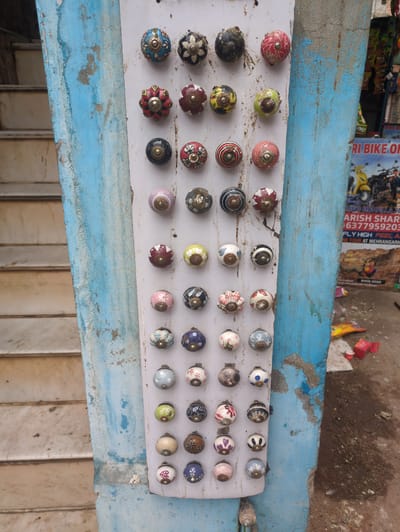 In Jodhpur, India, a display of decorative ceramic drawer knobs is mounted on a weathered, blue-painted pillar. The knobs, each approximately 2 inches in diameter, are arranged in a grid of six columns and seven rows. They feature a variety of designs, including floral patterns, geometric motifs, and solid colors such as red, blue, green, pink, and yellow, often with intricate metallic accents. Some knobs show signs of wear. To the left of the pillar, a staircase with light-colored steps is visible. To the right, a portion of a colorful advertisement for "RI BIKE ON" and "MEHRANGARH" is displayed, along with some scattered litter on the ground, including red boxes and yellow plastic packaging. The overall scene suggests a street-side stall in a bustling urban environment.