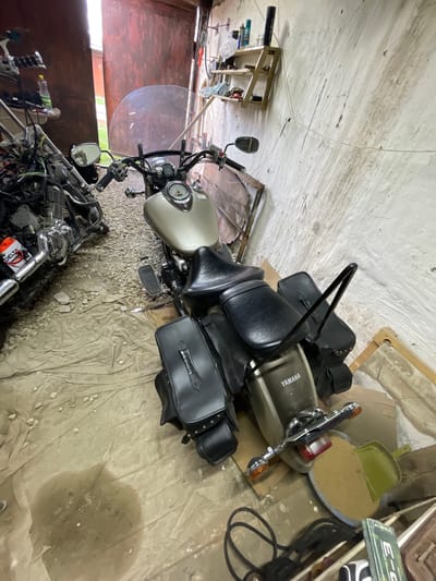 The image, rotated 90 degrees clockwise, depicts the interior of what appears to be a cluttered garage or workshop. The setting is in Znamensk, Russia, characterized by rough, light-colored walls showing signs of wear and dirt. The floor is mostly covered by a large, beige tarp or fabric, upon which gravel and debris are scattered, and a distinct wet stain is visible.
The primary subject is a cruiser-style motorcycle, light beige or tan in color, positioned centrally. The word "YAMAHA" is clearly visible on its fuel tank, which incorporates a speedometer. The motorcycle features black seats, black leather saddlebags with stud detailing, a passenger backrest, and a clear windshield. Chrome handlebars, mirrors, and other components add to its classic aesthetic.
A second motorcycle, also a cruiser, is partially visible in the bottom left, appearing to be in a state of disassembly or maintenance, with exposed engine components. A red and white battery labeled "DELTA" is identifiable on this bike.
Various other items populate the garage: wooden structures, possibly shelves or framing, are seen against the walls. Electrical wires run along the ceiling and walls. Near the rear of the main motorcycle, a green plastic dustpan holds sand in a blue tray. A green and white rectangular object with text that appears to be "E-C" or "Э-С" (Cyrillic characters) is also on the floor, alongside a coiled black cable. A plastic bottle is visible near the front of the second motorcycle.
No people are present. The scene suggests a storage area or a space where motorcycles are actively worked upon, under diffuse indoor lighting.