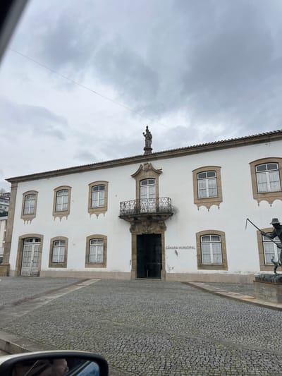 An exterior shot depicts the Câmara Municipal (Town Hall) of Santa Marta de Penaguião, Portugal, under an overcast sky. The prominent two-story building features a white painted facade with numerous rectangular windows trimmed in light-colored stone, each fitted with white frames and internal light-colored curtains. A central, ornate stone balcony with a black wrought-iron railing sits above the main entrance, which is dark and appears open. A stone statue of a robed figure, possibly religious, stands atop the roof directly above the balcony. The building's brown tile roof is visible. "CÂMARA MUNICIPAL" is inscribed in dark lettering on the white wall to the right of the main entrance. The foreground is a large cobblestone square. To the far right, a partial view of a bronze-colored statue on a stone pedestal shows a figure wearing a hat and holding a long-handled tool over his shoulder. In the lower-left corner, a car's side mirror is visible, reflecting a portion of the vehicle's interior and a person's face. Hills are discernible in the distant background on the left. A thin dark wire traverses the upper portion of the frame horizontally.