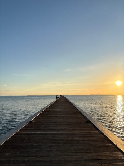 A long wooden pier extends into the ocean under a clear blue sky with a faint gradient of yellow and orange near the horizon. The setting sun is visible on the right side of the horizon, casting a golden reflection on the calm water. Two small figures are silhouetted on the pier in the distance, appearing to walk towards the setting sun. The pier is made of dark wood planks and has metal railings along its sides. In the distance, several islands are faintly visible on the horizon. The scene depicts a serene sunset over the ocean at Baan Taling Ngam, Thailand.