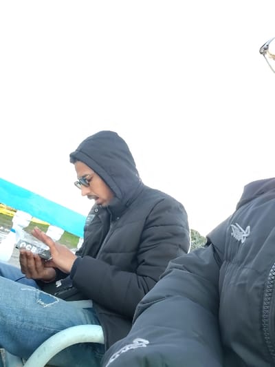 Two people are seated outdoors. The person on the left wears a black hooded puffer jacket, blue jeans with a ripped knee, and sunglasses. He holds a small rectangular object in his hands, examining it closely. The second person, positioned to the right, is partially visible. They are also wearing a black puffer jacket, with a white logo on the sleeve. The background is light, with a hint of blue and green.