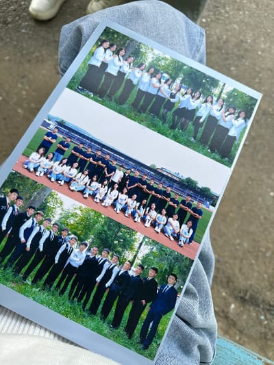 The image shows three photographs printed on a single sheet of paper, held by a person wearing grey denim jeans. The photographs appear to be of school groups.
The top photograph depicts a group of young women posing outdoors in what looks like a park or grassy area with trees in the background. They are all wearing similar white tops and dark skirts, with some adjusting their hair or clothing.
The middle photograph shows a larger group of young men and women posing in front of a stadium or sports field with bleachers visible in the background. The individuals are arranged in rows, some sitting on the ground and others standing. They are dressed in casual attire, including t-shirts and jeans.
The bottom photograph features a group of young men in formal attire, including suits and ties, posing outdoors in a grassy area with trees. They are arranged in multiple rows, with some individuals in the foreground wearing vests over their shirts and ties.
The overall setting for the photographs appears to be outdoors, possibly in a school or community setting. The context of the photographs suggests they are graduation or school event photos. The location mentioned, "Тайгараев, Kyrgyzstan," indicates the geographical context of these images, suggesting these events took place in that region. The person holding the photographs is dressed in casual denim, and the background suggests an outdoor urban or semi-urban environment with concrete and a worn wooden surface.