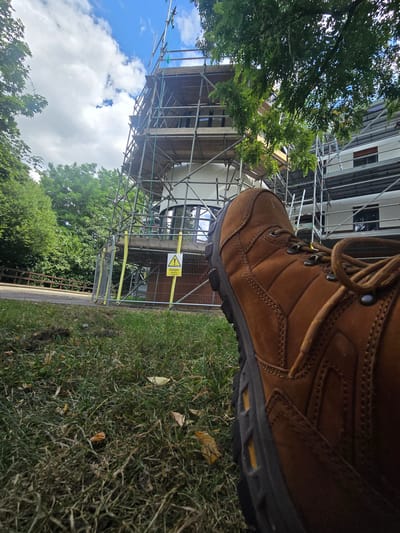 The image shows a close-up, low-angle view of a brown work boot in the foreground, resting on grass. Behind the boot, a building is under construction, covered in scaffolding. A yellow warning sign is visible on the scaffolding, indicating "Danger: Workers and Public." The environment appears to be outdoors, with trees and a blue sky with clouds visible. The scene is likely located in Woking, United Kingdom, given the context of construction and typical outdoor settings in that region.