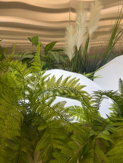 The image shows a close-up view of artificial plants, primarily ferns, in the foreground. Behind the ferns, there are white, curved surfaces that suggest a sculpted or mountainous terrain, possibly part of an interior design or a decorative display. In the background, feathery white plumes, resembling pampas grass, are visible along with some dark green, blade-like foliage. The backdrop appears to be a wall with a textured, wavy pattern in a golden-brown hue. The scene is illuminated, creating highlights and shadows on the plant materials and the white surfaces. No people or specific identifiable landmarks of Ladozhskoe Ozero, Russia are present in the image.