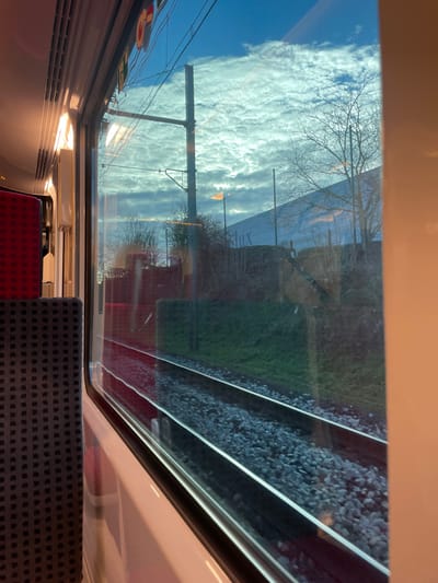 The image presents a view from inside a train carriage looking out a window. On the interior, a dark grey, patterned fabric seat is visible in the foreground to the left, with a red, patterned seat positioned behind it. Interior lighting fixtures emit a warm glow from the upper left, reflecting on the window glass.

Outside the train, a railway environment is depicted, located in Plaisir, France. Two parallel train tracks are visible in the lower half of the frame, resting on grey ballast stones. Adjacent to the tracks is a green grassy embankment. Beyond this, a sparse collection of leafless deciduous trees and bushes line the area. A tall, dark grey utility pole, supporting overhead electrical wires, stands prominently in the mid-ground. In the background, a large, light-colored building or wall, possibly a sound barrier, is partially visible, displaying the faint lettering "lux". A single, distant light source emits an orange glow. The sky above is partly cloudy, featuring numerous white, altocumulus-like clouds against a blue background. No individuals are present in the scene.