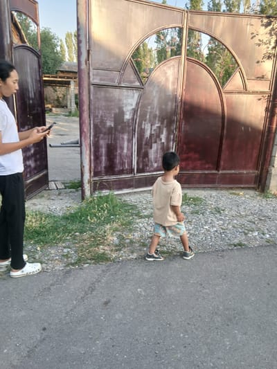 The image shows two people standing near a large, rustic metal gate in Тайгараев, Kyrgyzstan. On the left, a person is standing with their back to the camera, wearing a white t-shirt and black pants, and holding a black smartphone. White Crocs are visible on their feet. To the right, a young child, seen from behind, is walking away from the camera towards the gate. The child is wearing a light tan t-shirt and patterned shorts, with dark sneakers on their feet. The gate is made of dark metal with ornate circular and triangular patterns. Behind the gate, trees and buildings are visible. The ground in front of the gate is a mix of gravel and dry grass. The scene appears to be outdoors, possibly in a residential area, with a paved surface in the foreground.