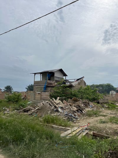 In Bundar, Indonesia, an overcast sky looms over a scene of destruction and disarray. The remains of a structure, likely a house, are partially visible amidst a landscape littered with debris. Piles of wood, corrugated metal sheets, plastic sheeting, and other construction materials are scattered haphazardly across the ground. Overgrown grass and vegetation are encroaching on the scene, suggesting a period of neglect or abandonment. The skeletal remains of the building, with its exposed wooden framework and partially intact roof, speak of a recent demolition or severe damage. The overall impression is one of a community in a state of rebuilding or recovery, with the remnants of past structures serving as stark reminders of past events. The presence of palm trees in the background hints at a tropical or subtropical environment.