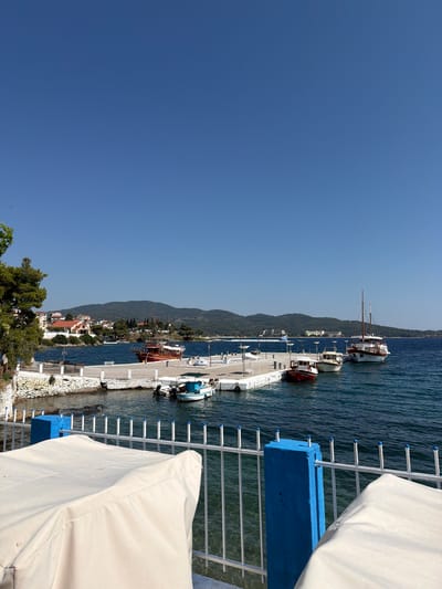 A bright, clear daytime scene depicts a harbor in the Sithonia Municipal Unit, Greece, with a focus on docked boats and the surrounding coastal landscape. The foreground features a white railing with blue decorative finials, separating a seating area with tables covered in white cloth from the water. Beyond the railing, a white pier juts out into the deep blue sea, where several boats are moored. A large, wooden sailing vessel with a prominent mast is docked on the right, next to two smaller fishing boats. A smaller, traditional red boat with a white roof is seen closer to the left side of the pier. Further back, more boats are visible, and the shoreline is lined with white buildings with red-tiled roofs, characteristic of a Greek coastal town. Green trees and lush vegetation grow along the hillside, which rises behind the town, eventually meeting the horizon with a range of distant mountains. The sky is a vibrant, cloudless blue, indicating fair weather.