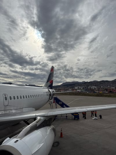 The image is taken from an elevated perspective, likely from inside an airplane cabin looking out onto the tarmac at Alejandro Velasco Astete International Airport in Wanchaq, Peru. A passenger jet is visible with its tail and parts of its wings and engines in view. A boarding ramp is connected to the plane, and several ground crew members wearing orange vests are standing on the tarmac. The city of Wanchaq is situated near a mountain range and is seen in the background under a heavily clouded sky, with a patch of sunlight shining through. An orange traffic cone is situated near the plane.