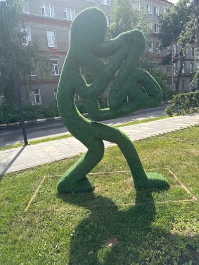 This image captures an outdoor sculpture made of artificial grass, resembling a stylized, abstract human figure in a seated, contemplative pose. The figure is composed of thick, rounded forms and appears to be intertwined with itself, with long limbs and a rounded torso. The sculpture is situated on a grassy area next to a paved walkway, with a street and a multi-story apartment building visible in the background.

The scene is bathed in sunlight, casting a distinct shadow of the sculpture onto the grass, suggesting it is likely daytime, possibly late morning or early afternoon given the angle of the sun and the length of the shadow. The weather appears clear and sunny. The apartment building is typical of Soviet-era architecture, with rows of windows and a grey brick facade, providing a clue to the urban or suburban setting. The presence of trees and other greenery suggests a park or residential area.

The overall mood is peaceful and perhaps a bit whimsical, with the artificial grass sculpture adding an artistic and unexpected element to the everyday setting of a residential street. There is no visible text in the image. The location context is Ramenskoye, Russia.