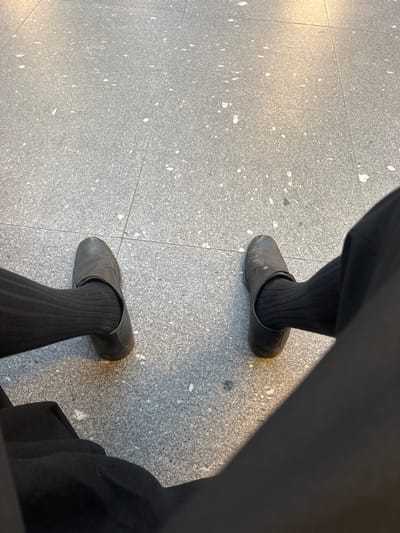 A low-angle, downward-looking image captures the lower extremities of one individual on a tiled floor. The person is wearing black trousers, black ribbed socks, and a pair of worn, dark-colored, slip-on shoes with visible scuff marks. The left foot is positioned slightly forward and to the left of the frame's center, while the right foot is to the right. The floor consists of light gray rectangular tiles with irregular white speckles, indicative of terrazzo or a similar composite material. Grout lines delineate the tile pattern. Reflections of overhead light sources are visible on the polished tile surface, particularly around the shoes. The individual appears to be seated or standing stationary. No specific identifying markers for a precise location within Guimarães, Portugal, are discernible from the visual information provided.