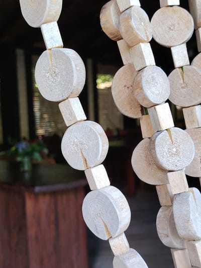 This is a close-up, eye-level, medium shot of a beaded curtain made of light-colored, natural wood. The curtain is composed of alternating round wooden discs and small cuboid wooden beads, strung together and hanging vertically. The round discs have a natural grain visible, and each one has a small, vertical cut in its surface. The cuboid beads are also made of wood and are slightly smaller than the discs. The curtain appears to be hanging in an entryway or as a room divider, as indicated by the blurred background. The background shows hints of a dimly lit interior space, with vertical blinds or window panes and a dark wooden structure, possibly furniture or a wall. There's also a glimpse of greenery and a purple flower in a vase on a dark surface in the mid-left of the background, suggesting an indoor setting with some decorative elements. The lighting is soft, and the time of day is not definitively clear, but the dim interior suggests it could be evening or a room with limited natural light. No people are visible, and no text is present in the image. The focus is entirely on the textured details and construction of the wooden beaded curtain.
