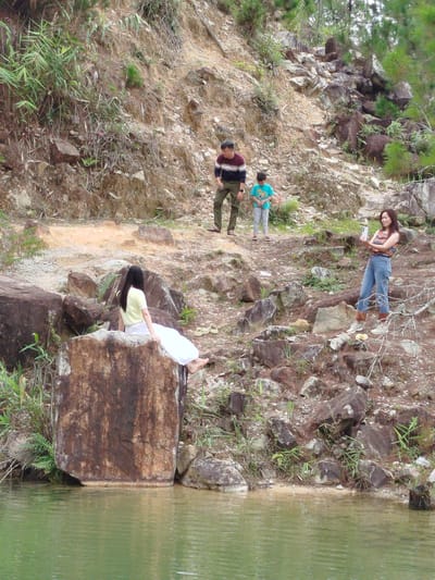 In the city of Lán Tranh, Vietnam, a group of four individuals are captured in a natural environment near a body of water. In the foreground, a woman with long dark hair, wearing a pale yellow top and white pants, is seated on a large, reddish-brown rock. Her back is to the camera, and her legs are extended towards the green water.
Further up a rocky, uneven slope, an adult male wearing a striped sweater and olive-green pants stands, facing the right. Beside him, a young child in a teal t-shirt and grey pants is looking down. On the right side of the image, a woman with dark hair, wearing a striped sleeveless top and blue jeans, is standing and holding a smartphone, appearing to take a picture or record a video. She is positioned higher up on the rocky terrain. The environment is characterized by loose soil, numerous rocks of varying sizes, and sparse vegetation, including grasses and some trees on the upper slopes. The water in the foreground is a murky green and appears still. The overall scene suggests an outdoor excursion or outing in a rural or natural setting within Lán Tranh.