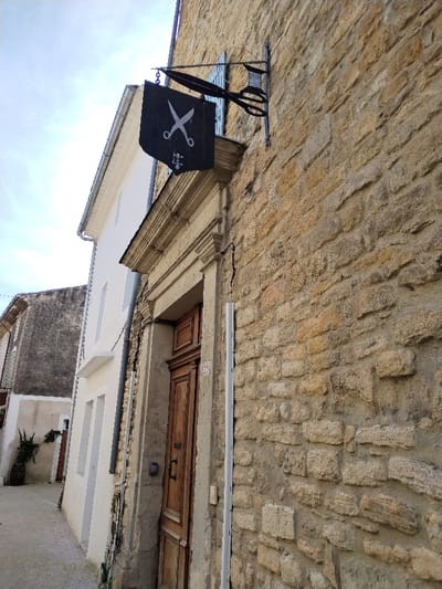 A stone building features a dark flag with white scissors hanging above an ornate doorway. The door is wooden with an address number visible. A metal sign with scissors extends from the building, suggesting a tailor or barber shop. A narrow street runs alongside the building, revealing a glimpse of another structure further down the street. The sky is partly cloudy.