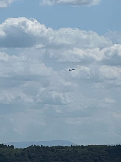 In the city of Karniowice, Poland, an airplane is seen flying in the sky. The sky is filled with clouds, and below the clouds, a dark green forest can be seen, with a line of distant hills on the horizon. The airplane is dark in color and is seen flying to the left of the image.