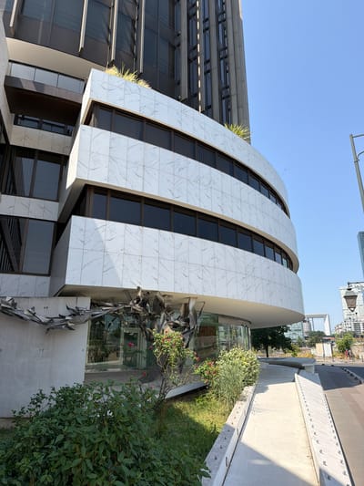 The image shows a curved building facade covered in white marble tiles with grey veining, set against a clear blue sky. The building has multiple levels of dark-tinted windows. In the foreground, lush green bushes and a flowering plant with pink roses are visible. A metallic sculpture extends from the left side of the building. To the right, a light-colored concrete ramp slopes upwards, bordered by metal posts. In the background, a tall modern building and other structures are present, suggesting an urban setting. The scene is captured on a sunny day. This building is the former headquarters of the oil company Elf Aquitaine, located in Neuilly-sur-Seine, France.