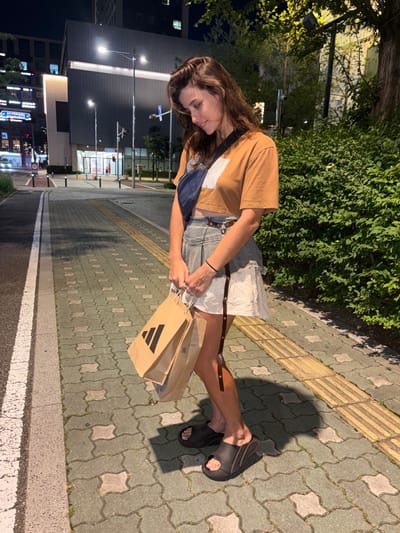 A woman with long brown hair and fair skin is standing on a sidewalk in Incheon, South Korea, at night. She is wearing a tan t-shirt, a denim skirt with white fabric underneath, and black slides. She is holding two brown paper shopping bags with the Adidas logo. The sidewalk is made of interlocking grey and green pavers with a yellow tactile warning strip. Behind her, a modern building with illuminated windows and streetlights can be seen. Trees and bushes line the right side of the image. The street is to the left, with streetlights and traffic signals visible.