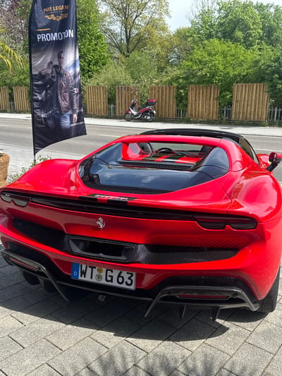 The image shows a red Ferrari parked on a paved surface in Jestetten, Germany. The car's rear is prominently displayed, including the license plate reading "WT G 63". A black banner with the words "PME LEGEND AMERICAN CLASSIC" and "PROMOTION" is visible to the left of the car. In the background, a wooden fence, green trees, and a parked scooter can be seen. The scene appears to be outdoors, likely on a street or parking area.