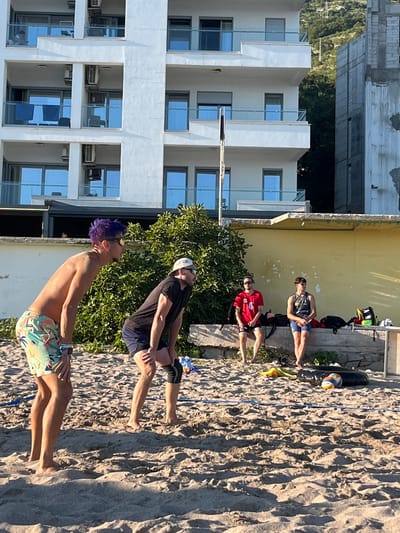 In Miljevci, Montenegro, on a sandy beach, two young men are playing beach volleyball. The man in the foreground on the left has purple hair and is wearing colorful shorts and sunglasses. The man next to him on the right is wearing a black t-shirt, dark shorts, and a knee brace, and also sunglasses. In the background, two other individuals are seated on a stone ledge. One is wearing a red t-shirt with the number "4" and black shorts, while the other is in a grey tank top and blue shorts. They are all facing the volleyball court, which is partially visible with a net and two balls. Behind them, a modern white building with multiple balconies stands next to a construction site. Lush green trees and a mountain are visible in the distance. The lighting suggests it is late afternoon.