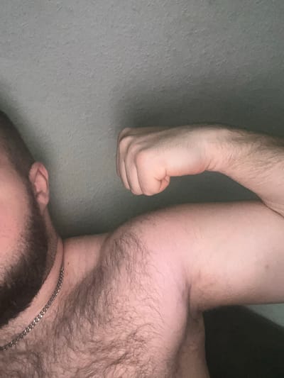 This is a close-up, low-angle photograph of a person's upper body, likely taken indoors. The subject's left arm is flexed, with the fist clenched. The skin is light-toned and appears to be a male. The subject has a beard and chest hair, and is wearing a silver chain necklace. The background is a textured, light gray wall. There is no indication of the city of Gelsenkirchen, Germany, or any other specific location. The image is dimly lit, with the light source appearing to be from the upper right.