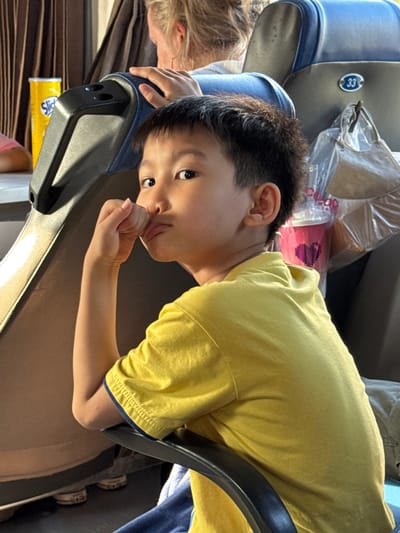 The image depicts a young East Asian boy, approximately 5-7 years old, seated on a bus. He is wearing a bright yellow t-shirt and has dark hair. The boy is looking directly at the camera with a thoughtful expression, his right hand near his mouth. He is positioned in the foreground, with the background showing bus seats and curtains. To the left, a yellow canister with a "Slick" label is visible. To the right, a pink cup with a heart design is partially obscured by a clear plastic bag containing a mask. A seat number "33" is affixed to the seat behind the boy. The ambient light suggests it is daytime. The scene appears to be inside a passenger bus. There is no specific indication of the location being Tuy Hoa, Vietnam.