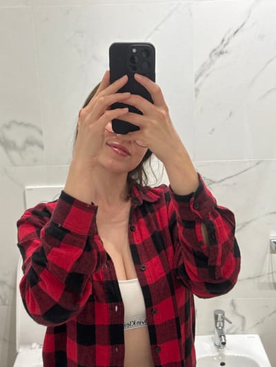 A mirror selfie depicts an individual within a private bathroom environment in Scafati, Italy. The person, positioned centrally, holds a black smartphone with both hands, obscuring the individual's upper face. The visible facial features include the nose, glossy lips, and chin, with portions of eyeglasses visible. Brown hair frames the visible part of the face. The individual is wearing an unbuttoned red and black plaid flannel shirt, revealing a white athletic bra with an inverted "Calvin Klein" logo on its band. Visible cleavage is present.

The mirrored background features white tiles with grey marble veining. In the lower right of the reflection, a white ceramic basin with a chrome single-lever faucet is present. A white rectangular fixture, consistent with a toilet cistern, is partially visible to the left of the basin. A small chrome fixture is mounted on the wall to the right, above the basin.