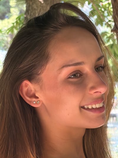 A young woman with light brown hair is smiling and looking upward. She has fair skin, light-colored eyes, and is wearing earrings in her left ear. The background features foliage, suggesting an outdoor setting. There is no clear indication of the location being Bunyola, Spain, from the provided image.