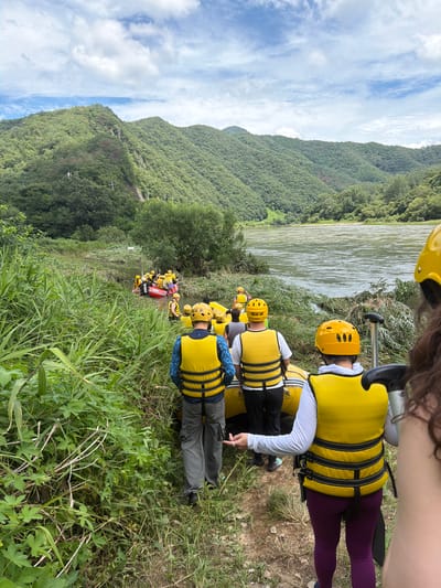 In the Seo-myeon area of South Korea, a group of people are preparing for a rafting excursion. The scene is set by a wide, calm river flowing between lush, green, tree-covered mountains under a sky dotted with white clouds. On the riverbank, amidst tall grasses and foliage, several individuals are visible, predominantly wearing bright yellow life vests and helmets. In the foreground, a person in a yellow vest and grey pants faces away from the camera, gesturing with their left hand. To their right, another person, also in a yellow vest and white long-sleeved shirt, is partially visible. Further into the scene, a line of people in yellow vests and helmets are walking towards and preparing to board inflatable rafts in the shallow water. The rafts are yellow and red, and some are already partially submerged. The environment is natural and undeveloped, with the river and mountains forming a serene backdrop for the recreational activity.