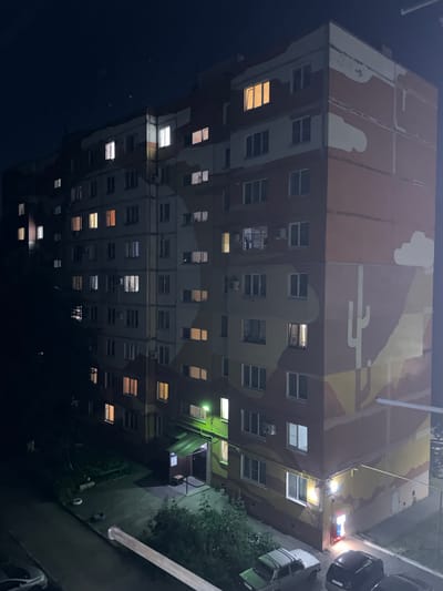 This is a night-time, high-angle shot of a multi-story apartment building located in the city of Taganrog City District, Russia. The building is dark, with some windows illuminated from within, indicating occupied apartments. The exterior of the building features a mural with a desert landscape theme, including a stylized cactus. In the foreground, a paved area with some greenery, benches, and parked cars is visible. A street light illuminates a section of the ground near the building's entrance. The sky is dark and appears clear.