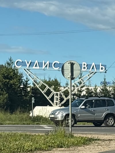 The image shows a sign with white Cyrillic lettering that reads "СУДИСЛАВЛЬ" against a blue sky with scattered clouds. The sign is mounted on a white metal structure that resembles an archway. In front of the sign, a silver SUV is driving on a road. The area around the road is covered in green grass and trees. The sign is likely a welcome sign for the city of Sudislavl, located in the Western Oblast of Russia.