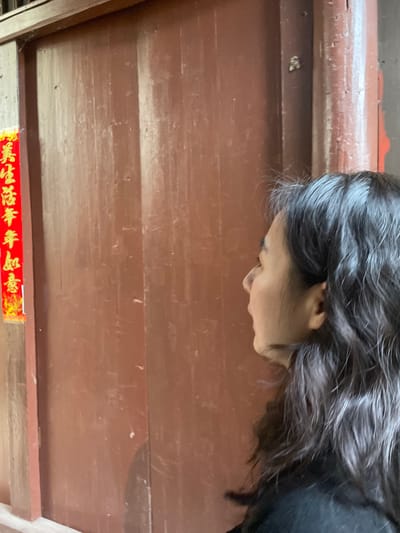 A woman with dark hair bends over a brown, wooden surface. The surface has horizontal planks, and a red and gold decorative banner is visible at the bottom. The woman's head is angled downwards, obscuring her eyes but showing her nose, mouth, and ear. Her hair is thick and slightly wavy. She appears to be looking at something on or below the wooden surface.