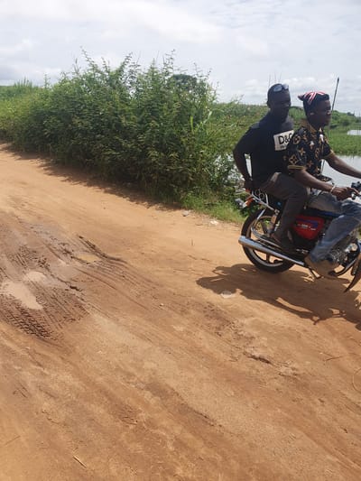 Two men are depicted on a motorcycle on a dirt road in Idah, Nigeria, under a cloudy sky. The image is taken from a slightly low angle, showing the road, lush green vegetation, and the sky.
The man driving is wearing a black t-shirt with "D&G" prominently displayed, and "DOLCE&GABBANA" in smaller text beneath it. He also wears sunglasses. The passenger behind him is dressed in a patterned shirt, possibly with a luxury brand motif, and a distinctive red, white, and blue patterned cap or headwear. The men appear to be mid-journey, possibly paused or moving slowly along the unpaved path.
The motorcycle is a standard model, with visible chrome exhaust pipes, wheels, and parts of its engine and frame. A red section, likely the fuel tank, is partially visible. The shadows cast on the ground suggest it is daytime, with the sun obscured by clouds, leading to softer lighting.
The setting is an outdoor, possibly rural or semi-rural area. The road is made of reddish-brown dirt, showing numerous tire tracks and some muddy puddles, indicating recent moisture. To the left side of the road, thick, tall green bushes and vegetation grow, hinting at a natural environment, potentially bordering a body of water visible in the distant background. The sky is largely overcast with white and grey clouds.