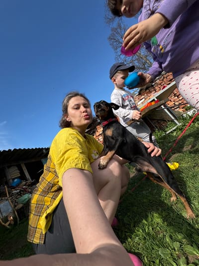 The image shows a group of people and a dog outdoors. A woman in a yellow shirt and plaid vest is holding a black and tan dog on her lap. Two children are partially visible above, holding blue and pink plastic Easter eggs. Behind them is a stack of bricks. The sky is blue. The location cannot be verified with the available information.