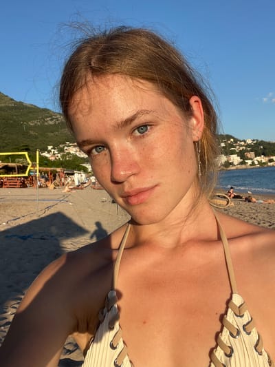 The image is a close-up selfie taken on a beach in Miljevci, Montenegro. The subject, a young woman with light brown hair and blue eyes, is wearing a beige bikini. She has freckles across her nose and cheeks. Her hair is slightly windswept. In the background, the beach is visible with scattered beach chairs and umbrellas, a volleyball net, and the sea with buildings on the hillsides. The sky is clear and blue, indicating it is daytime with bright sunlight, casting shadows on the sand. There are other people visible in the distance on the beach and in the water.