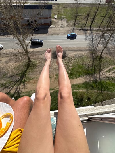 The image captures a top-down view from a balcony or high window in Ново-Никольское, Russia, showing two individuals relaxing while overlooking a street scene below.
In the foreground, a pair of bare, tanned legs, likely belonging to a female, are stretched out, with painted toenails visible on the feet. Adjacent to these, a portion of another person's leg and knee is seen, wearing white shorts, suggesting a male. A white, textured plate with orange peels rests near this person, beside a yellow cloth or garment. A blue patterned mat is partially visible beneath one of the legs, contributing to the relaxed atmosphere.
The view beyond the balcony railing reveals a residential or commercial street. Two cars are parked or driving on the asphalt road: a distinct blue sedan and a dark-colored car, possibly an SUV or another sedan. A wide grassy verge with scattered patches of dirt separates the road from a row of bare-branched trees, which cast long shadows, indicating a sunny day.
On the left side of the street, a multi-story building is visible, with a prominent sign on its side that clearly reads "WILDBERRIES," identifying it as a store or distribution center for the Russian e-commerce giant. Further down the street, another smaller building or structure is partially seen. A circular road sign with a red border and the number "80" inside, likely indicating an 80 km/h speed limit, is visible on a pole near the street.
The overall scene suggests a pleasant, sunny day, likely midday or early afternoon given the shadows, with people enjoying a leisurely moment outdoors on a balcony.