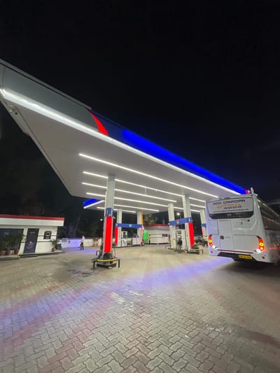 At a brightly lit petrol station in Kajheri, India, at night, a white bus with "INDO CANADIAN EXPRESS" written on its back is parked under a large canopy. The canopy is illuminated with white strip lights and accented with red and blue neon lights. The ground is paved with interlocking concrete blocks, reflecting the colored lights. To the left, a small building with "OFFICE" signage is visible, with potted plants outside. Several people are present, including a man standing near the office and others tending to the fuel pumps. A few motorcycles are also parked nearby. The overall scene depicts a busy, well-lit service station operating after dark.