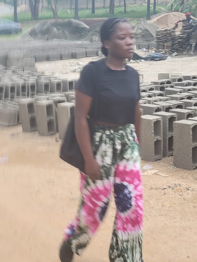 An outdoor, slightly blurry photograph, taken during the daytime in Kwamba, Nigeria, shows a young woman standing amidst a scene dominated by concrete blocks. The image is oriented vertically (originally rotated 90 degrees counter-clockwise).
The central subject is a woman, likely in her late teens or early twenties, with dark skin and braided or dreadlocked hair. She is wearing a plain black t-shirt and vibrant, tie-dye patterned trousers featuring shades of pink, green, white, and dark blue. A black bag is slung over her left shoulder. She is looking slightly to her left, with a neutral expression.
The ground she stands on is unpaved, appearing to be dirt or sand. To her right and behind her, numerous grey concrete blocks (also known as cinder blocks) are stacked in organized piles, suggesting a construction site or a block-making facility. In the mid-ground and background, more blocks are visible, along with a large mound of earth or sand. Faintly in the distance, another person can be seen working or moving near more stacks of materials and what appears to be part of a structure or machine. Green foliage and tree trunks are visible on the left side of the frame, indicating an outdoor, somewhat natural setting. A dark-colored water bottle is partially visible on the ground near the left edge.
The overall scene depicts an active work environment related to building materials or construction, with the woman perhaps observing or participating. No discernible text is visible in the image. The lighting suggests an overcast day or indirect sunlight.