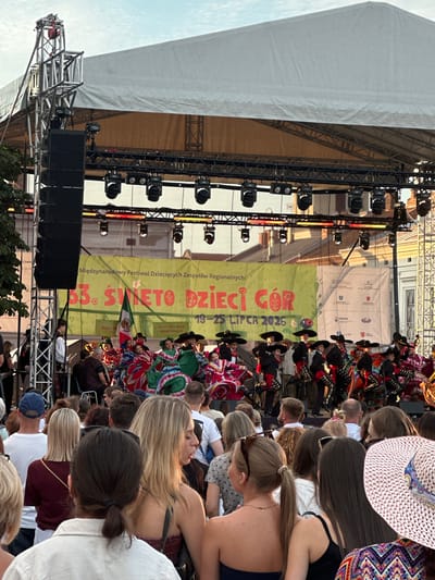 The image depicts an outdoor event on a clear day, likely late afternoon or early evening, with a bright sky visible. In the foreground, a crowd of people are gathered, their backs mostly to the camera, indicating they are facing the stage. They appear to be spectators at a performance.
The main focus of the image is a stage set up for a performance. A large, light-colored canopy covers the stage and a significant portion of the rigging above. Below the canopy, a large yellow banner is displayed, featuring text in Polish and a graphic of flowers. The text on the banner reads "Międzynarodowy Festiwal Dziecięcych Zespołów Regionalnych 33. ŚWIĘTO DZIECI GÓR 19-25 LIPCA 2026". This indicates the event is the "33rd Festival of Regional Children's Groups" and it took place between July 19-25, 2026. A Mexican flag is prominently displayed on the stage.
On the stage, a group of performers, dressed in traditional Mexican attire including sombreros and brightly colored dresses, are engaged in what appears to be a dance performance. Some performers are holding musical instruments. The stage is equipped with lighting rigs and large speakers are visible to the left of the stage, mounted on scaffolding. The overall setting suggests a cultural festival or celebration.
The image does not contain any visual cues that definitively place it in the city of Nowy Sącz, Poland, other than the Polish text on the banner.
