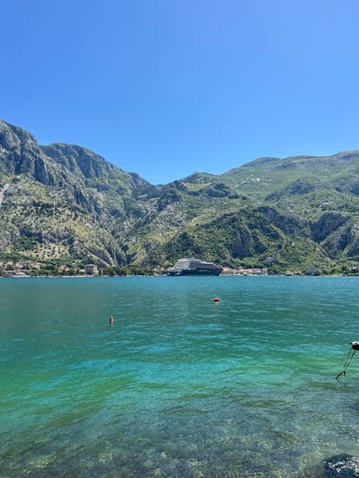 The photograph presents a view in Prčanj, Montenegro, featuring a mountainous landscape alongside the sea. The left side of the image showcases a rugged mountain range with dense vegetation. Nestled between the mountain and the shoreline are buildings, indicative of residential or commercial structures.
The right side depicts the sea, with varying shades of green and blue suggesting depth and differing light penetration. Two visible orange buoys float on the surface of the water. In the upper-right corner, there are fishing rods placed on the shore. The sky is clear blue, contrasting with the landscape below.