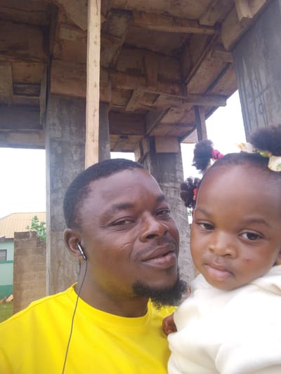 This is a medium close-up, eye-level selfie featuring an adult man holding a young child, both looking directly at the camera. The man, on the left, has dark skin, short dark hair, and a dark, trimmed beard. Distinct facial scars are visible on his left cheek (viewer's right). He wears a bright yellow t-shirt and has a black earphone in his left ear, bearing the text "CHAMPION" and "C-100". He exhibits a slight, pleasant smile.

The child, a toddler, is held on the right side of the frame. She also has dark skin and her curly dark hair is styled into two puffs, adorned with colorful, patterned hair ties (pink and yellow/white). She is dressed in a cream or white hooded top and has a neutral, observant expression.

The background reveals an outdoor setting, likely within or under a structure that appears to be under construction or unfinished. Large concrete pillars and raw wooden beams and planks forming a ceiling or roof are visible above and behind the subjects. Through gaps in the structure, a bright, overcast sky is seen, indicating daytime. To the far left in the background, a simple building with a green lower section and a lighter roof, along with some foliage, suggests a community setting in Dungu, Ghana.