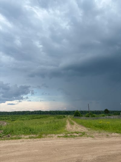 The image depicts a rural landscape under a stormy sky. In the foreground, a dirt road curves through a grassy field. In the middle ground, a line of trees forms a dense forest. In the background, the sky is dominated by dark, ominous clouds, suggesting an approaching storm. Power lines and poles are visible against the sky, indicating the presence of human infrastructure. The overall impression is one of an impending weather event in a natural setting. The location is identified as the city of Kurgan, Russia. There are no people or animals visible in the scene. The environment appears to be a field and forest, with a dirt road and utility poles.