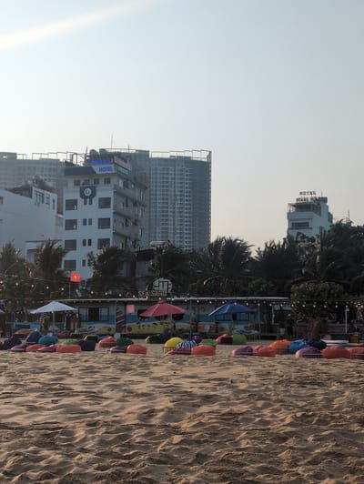 The image shows a beach scene in Quy Nhon Ward, Vietnam. The foreground is dominated by a wide expanse of sand. A line of colorful beach umbrellas and beanbag chairs is arranged along the edge of the beach. In the background, several buildings are visible, including hotels. One building is labeled "SEA SANDS." Another building is labeled "HOANG NHU HOTEL." A Vietnamese flag is flying in the middle ground. The sky is clear and light.