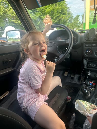 The image shows two people inside a car. The primary subject is a young girl with blonde hair, wearing a pink and white striped t-shirt and shorts. She is sitting in the passenger seat, looking towards the camera with a wide smile and her tongue sticking out, holding a lollipop. Her legs are extended, resting on the seat. Behind her and slightly out of focus, another person, likely an adult, is visible in the driver's seat, with their hands on the steering wheel. The interior of the car is visible, including the dashboard, steering wheel, gear shift, and pedals. The exterior of the car is reflected in the side mirror and visible through the windows, showing green trees and a bright, sunny sky, suggesting it is daytime. A sign with Russian text is visible through the windshield, with the location context identifying the setting as Кварса, Russia. The overall mood appears cheerful and informal, possibly indicating a casual outing or travel.