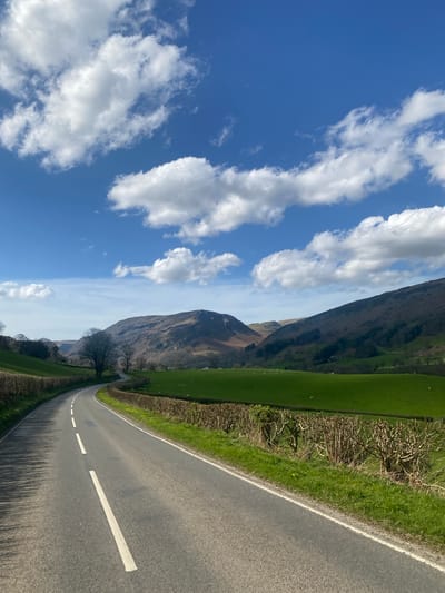 The image depicts a rural road scene in the Pen-y-bont-fawr area of the United Kingdom. The road, marked with a central broken white line, curves to the left. A grass verge and a hedgerow line the right side of the road. Beyond the hedgerow is a field with a gradual incline towards a hill. The sky is blue with scattered white clouds. A tree is on the left side of the road.