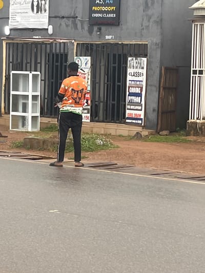 A person, seen from the back, stands on a street in Sabonjibi, Nigeria. They are wearing an orange t-shirt with a graphic print and black track pants with white stripes. The person is also wearing black flip-flops. The scene appears to be in front of a building with barred windows and several signs.

One sign, mounted above the barred windows, advertises "CODING CLASSES" with specific mentions of "PYTHON" and "COREL DRAW". Another sign to the left, also in portrait orientation, reads "A3, A4 PHOTOCOPY CODING CLASSES" and "SHOP 1". A white banner with the text "Rosy Hair" is visible on the far left.

The ground is a mix of paved road and dirt with some sparse vegetation. The lighting suggests it might be daytime, possibly overcast or late afternoon, as there are no strong shadows. The overall impression is that of a commercial street in Nigeria.