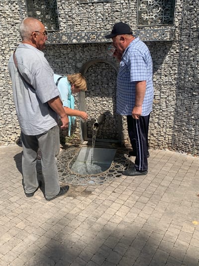 In the city of Arbuzov-Baran, Russia, three individuals are gathered near a public water fountain. The fountain is integrated into a wall constructed of grey stones, with decorative metal elements above. A metal grate covers a pool beneath the spout of the fountain, from which water is flowing.

On the left, an older man with short grey hair and glasses stands facing away from the viewer, wearing a light grey short-sleeved collared shirt and grey trousers. A brown strap is slung across his shoulder.

In the center, a woman with short blonde hair is leaning over the fountain, her back mostly to the viewer. She is wearing a light blue top and appears to be interacting with the water flow.

On the right, another older man, wearing a blue and white plaid short-sleeved shirt, dark trousers with white stripes, and a black baseball cap, is drinking from a clear plastic cup. He is facing towards the fountain.

The ground is paved with light-colored rectangular stones. The overall scene suggests a sunny day, with shadows cast by the people and the wall.