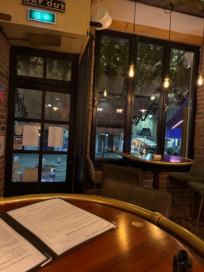 A bar interior shows a round table with a brass edge and menus. Two chairs with green upholstery are visible. The scene is lit by vintage-style light bulbs strung across a multi-pane window, offering a glimpse of the outside. An illuminated "WAY OUT" sign is on the wall. Brickwork frames the windows and walls.