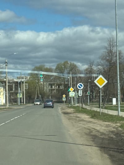 The image shows a street scene in Ново-Никольское, Russia, with a view of a road running alongside a grassy area. Several traffic signs are visible along the roadside, including a yellow diamond-shaped sign indicating a hazard, a blue and white sign indicating "no entry," and other directional and informational signs. Traffic lights are also present. Two cars are seen driving on the road. The sky is visible in the background, showing a mix of blue sky and clouds. The image appears to have been taken from a vehicle.