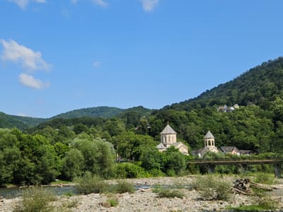 In Plastunka, Russia, a tranquil river flows through a picturesque landscape, bordered by a rocky embankment and lush green trees. On the opposite bank, a charming village nestles amidst the verdant hills. Two prominent buildings, possibly churches, stand out with their distinctive architectural styles, adorned with domed roofs and bell towers. A weathered bridge spans the river, connecting the village to the other side. The sky above is a vibrant blue, with a few scattered clouds adding to the serene atmosphere. The scene evokes a sense of peace and natural beauty.