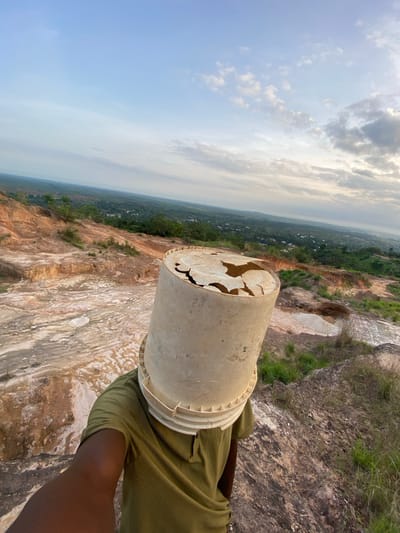 The image presents an outdoor scene from a high vantage point, likely a cliff or quarry, in Ufueseni, Nigeria. The primary subject is a partially visible person, seen from a high angle, whose right arm and torso, clad in an olive-green ribbed shirt, are visible on the right side of the frame. A large, light-colored (beige or off-white) plastic bucket with a severely broken and jagged bottom is balanced on the person's head. The inside of the bucket, showing its cracked structure, is clearly visible.
The setting is characterized by a dramatic landscape. Immediately below and to the left of the person is a steep, rugged cliff face, displaying exposed rock in various earthy tones, from light sandy yellows to darker browns and greys. Patches of green grass and other vegetation cling to the rock, particularly on flatter areas and along the top edge of the cliff.
In the mid-ground and distant background, the terrain opens up to reveal a vast expanse of rolling hills covered in dense green foliage, suggesting forests or agricultural land. Scattered small, light-colored structures can be faintly seen within the distant green landscape, indicating a remote settlement or village. The sky above is bright, with a mix of clear blue areas and scattered white and grey clouds, consistent with daytime and fair weather conditions. The overall impression is one of immense natural scale and a striking contrast between the rugged foreground and the lush distant landscape. No visible text is present in the image.